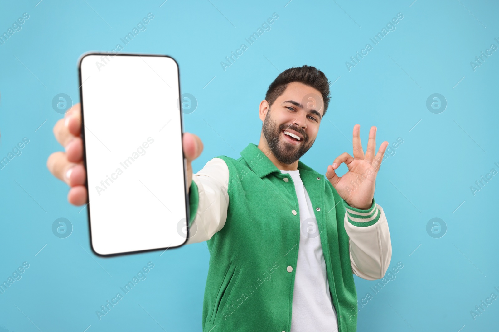 Photo of Young man showing smartphone in hand and OK gesture on light blue background