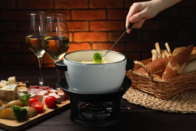 Photo of Woman dipping piece of broccoli into fondue pot with melted cheese at wooden table with wine and snacks, closeup