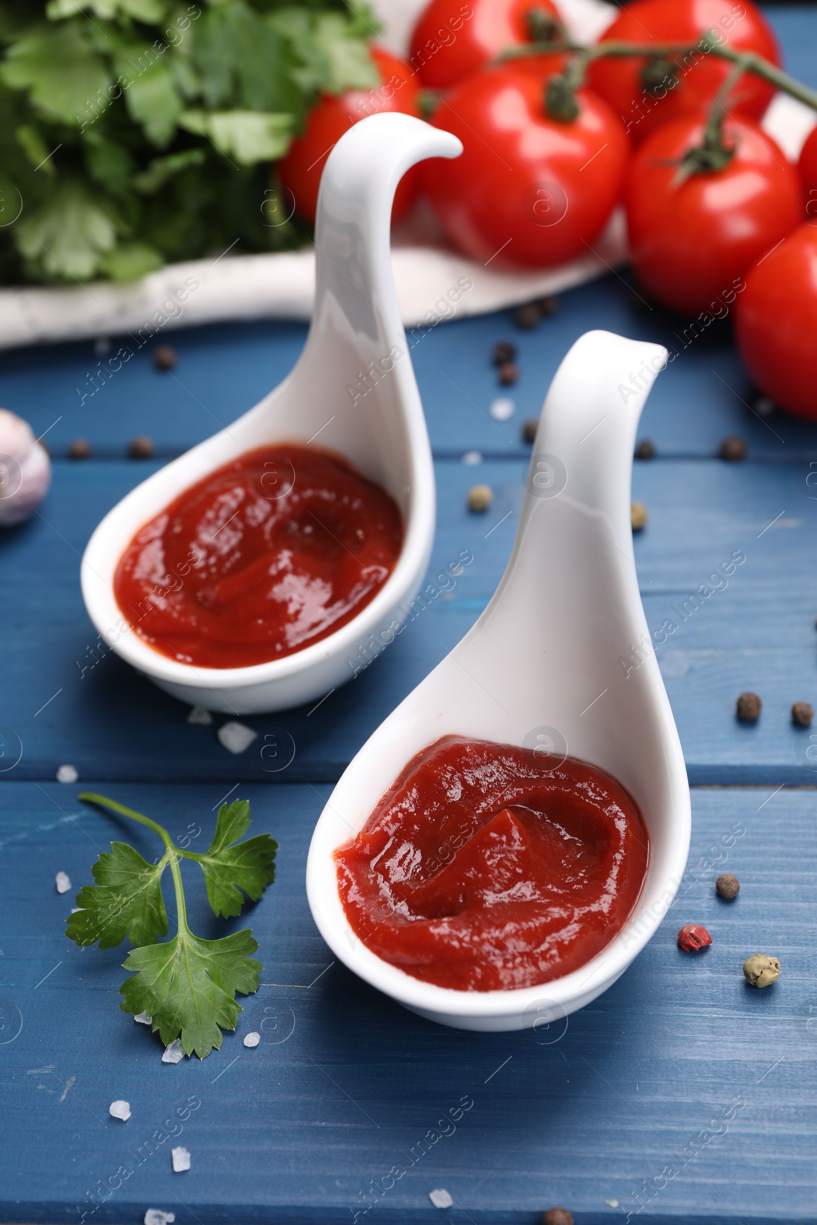 Photo of Organic ketchup in spoons, fresh tomatoes and spices on blue wooden table, closeup. Tomato sauce