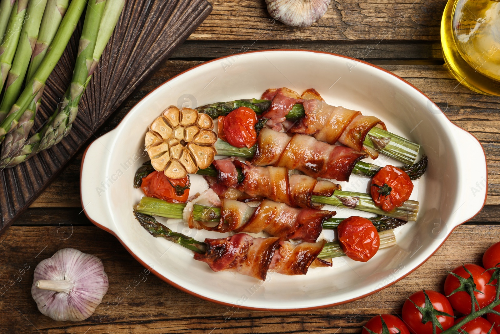 Photo of Oven baked asparagus wrapped with bacon in ceramic dish on wooden table, flat lay