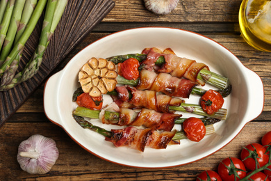 Oven baked asparagus wrapped with bacon in ceramic dish on wooden table, flat lay