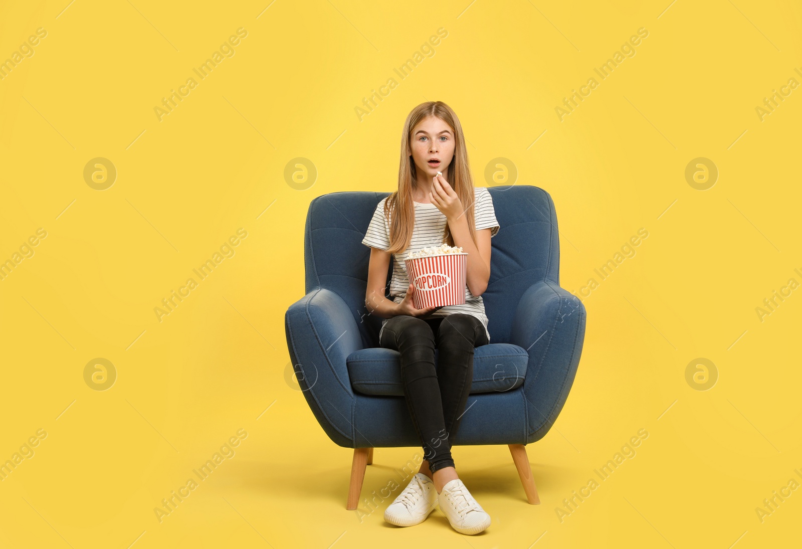 Photo of Emotional teenage girl with popcorn sitting in armchair during cinema show on color background