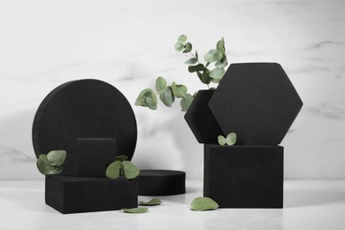Photo of Black geometric figures and eucalyptus leaves on white marble table. Stylish presentation for product