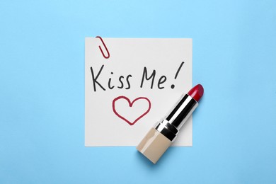 Photo of Card with phrase Kiss Me and drawn heart, lipstick on light blue background, top view