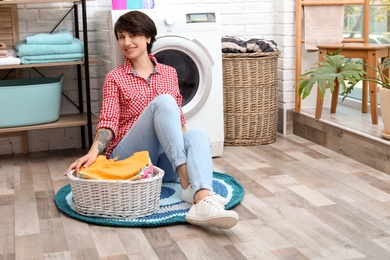 Photo of Young woman with basket full of clean clothes near washing machine at home
