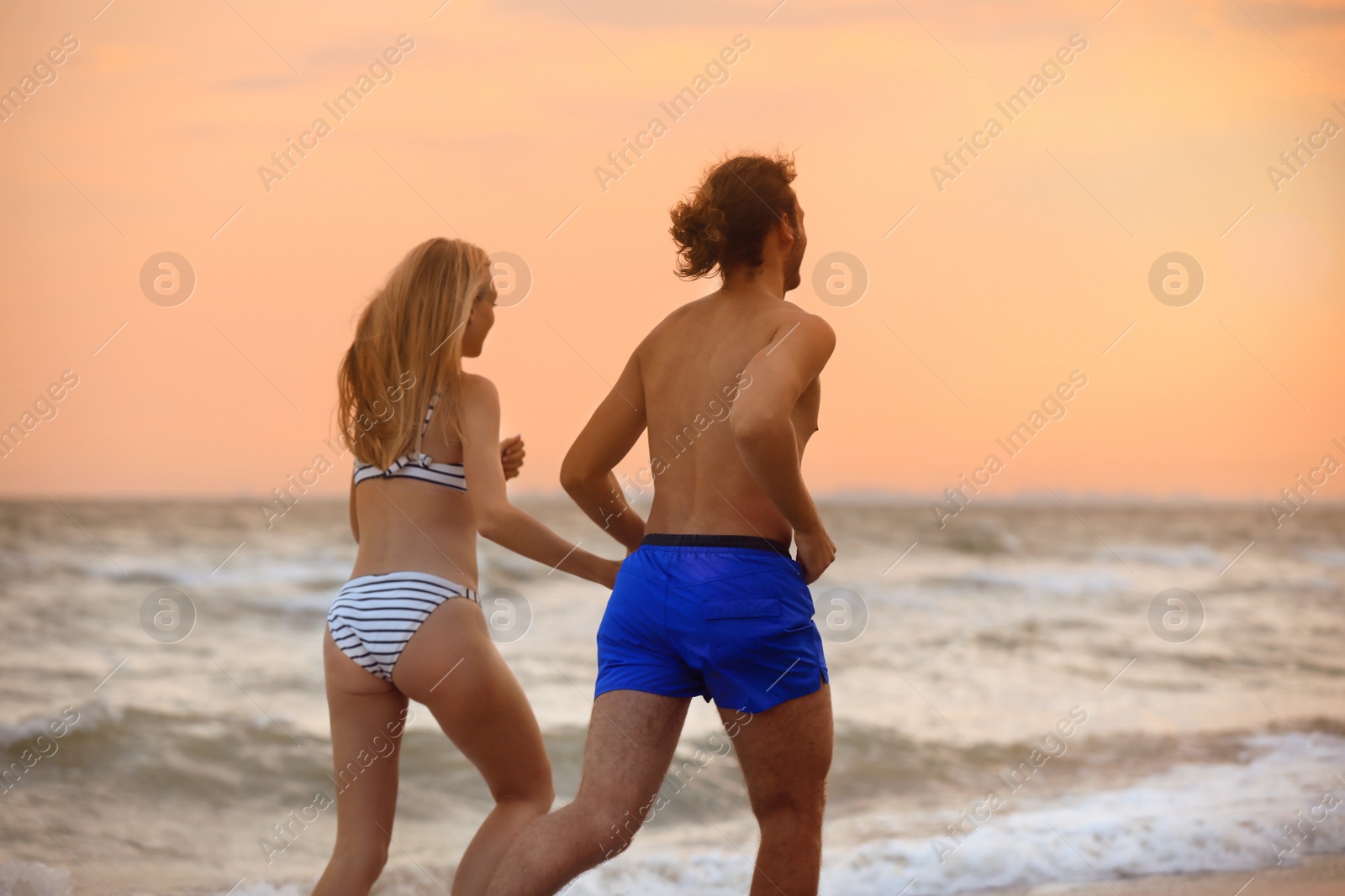 Photo of Young woman in bikini and her boyfriend walking on beach at sunset. Lovely couple