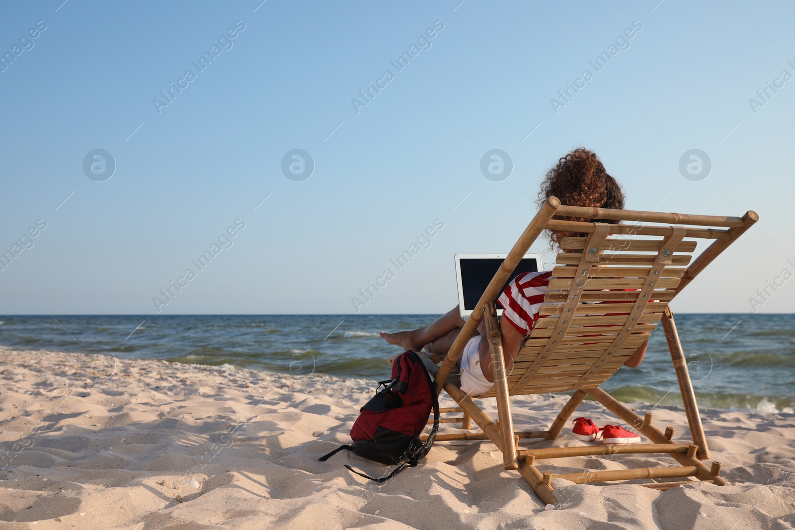 Photo of African American woman working on laptop in sunbed at beach, back view