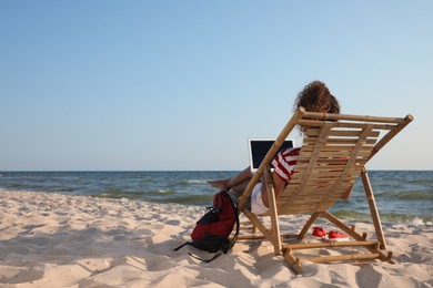 African American woman working on laptop in sunbed at beach, back view