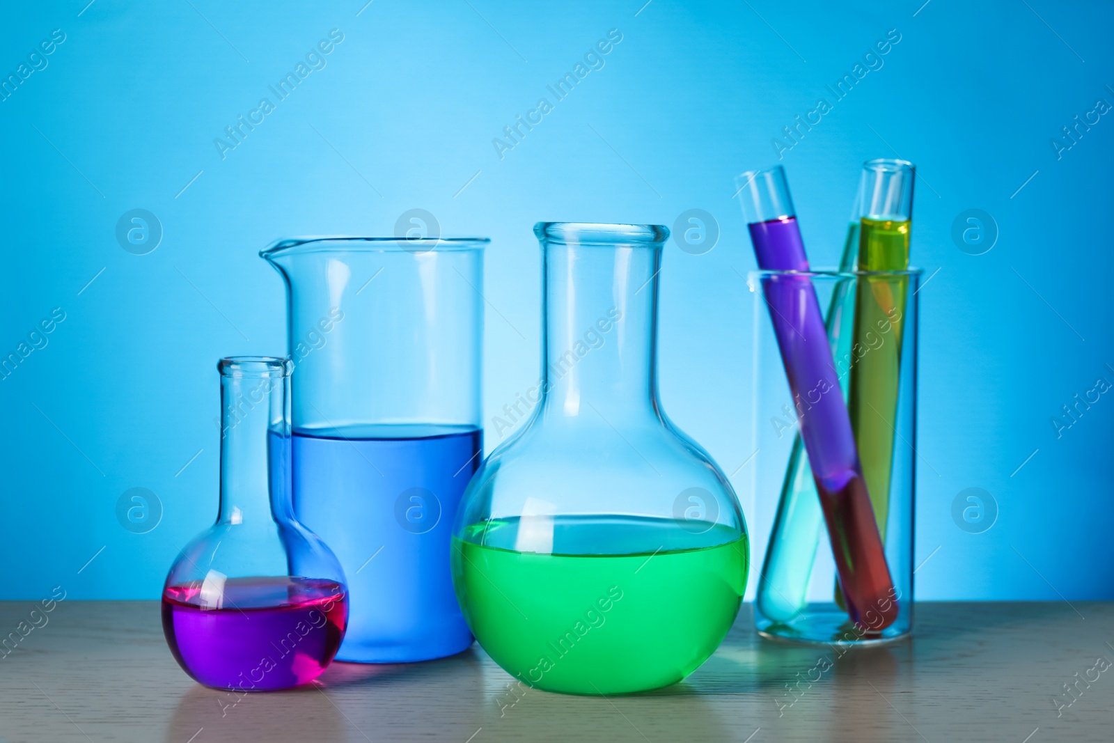 Photo of Different laboratory glassware with colorful liquids on wooden table against light blue background