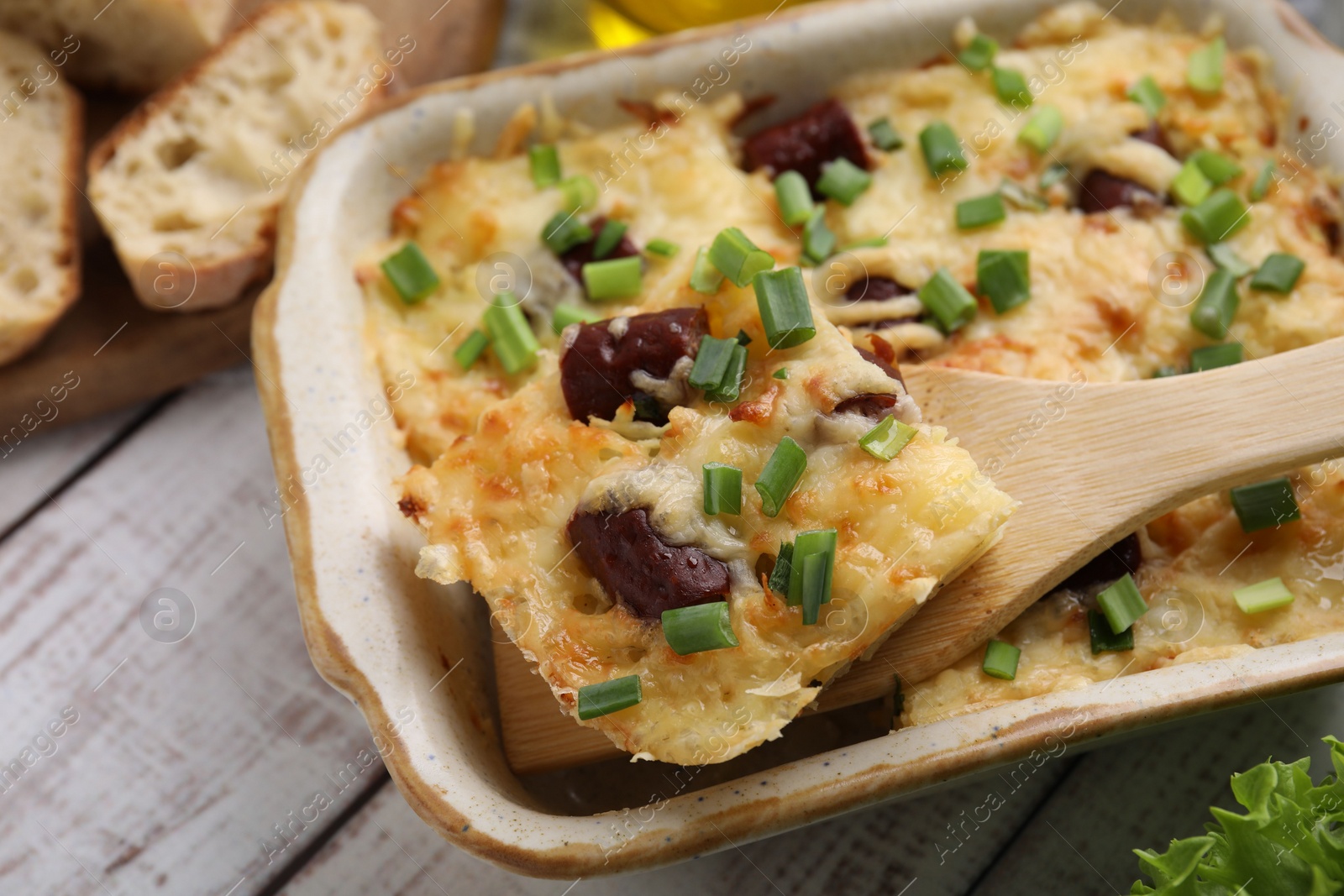 Photo of Taking piece of tasty sausage casserole from baking dish at white wooden table, closeup