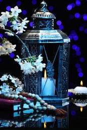 Photo of Arabic lantern, Quran, misbaha, burning candle and flowers on mirror surface against blurred lights at night