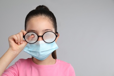 Photo of Little girl wiping foggy glasses caused by wearing medical face mask on grey background, space for text. Protective measure during coronavirus pandemic