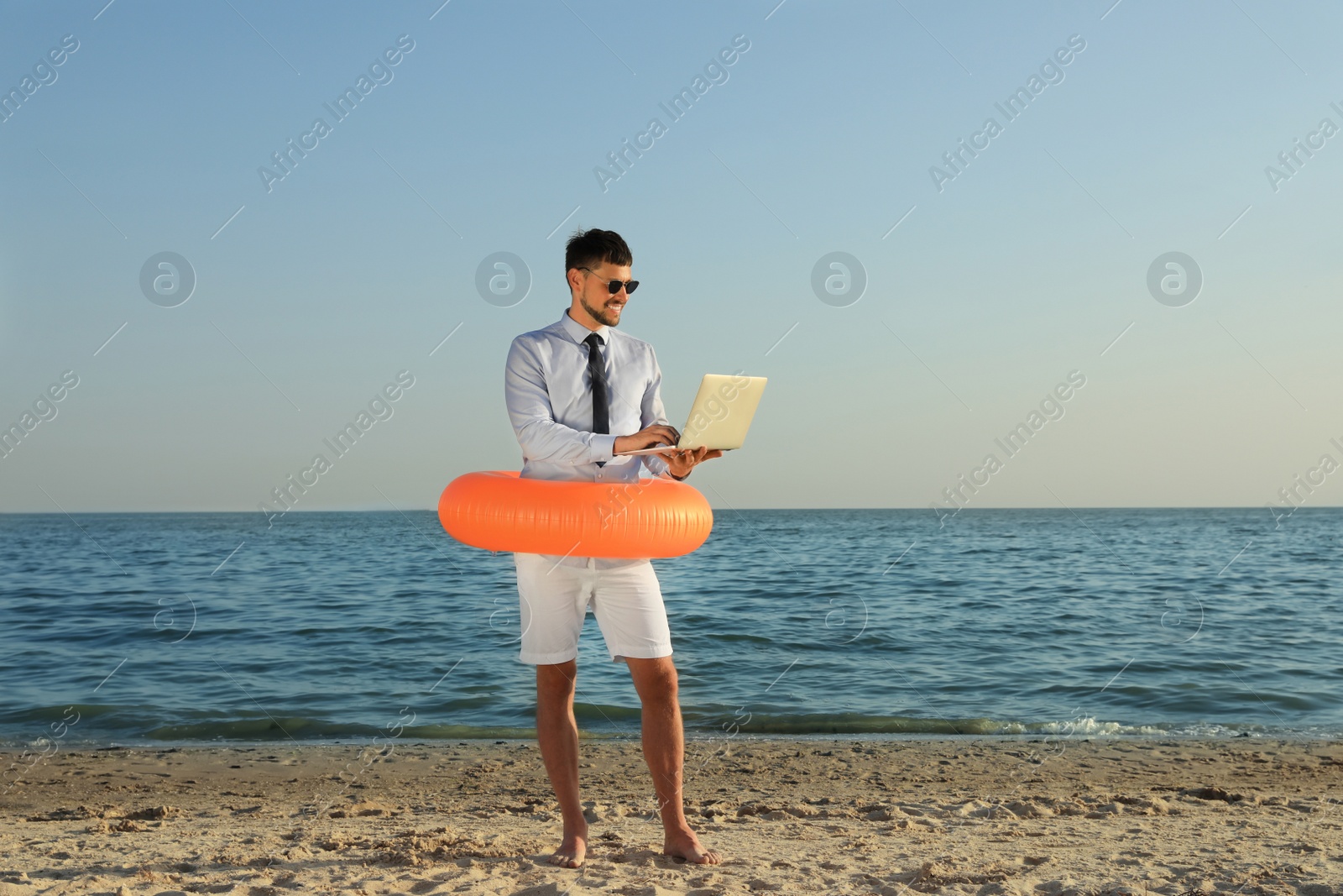 Photo of Happy man with inflatable ring and laptop near sea on beach, space for text. Business trip