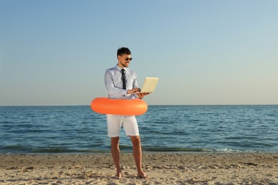 Happy man with inflatable ring and laptop near sea on beach, space for text. Business trip
