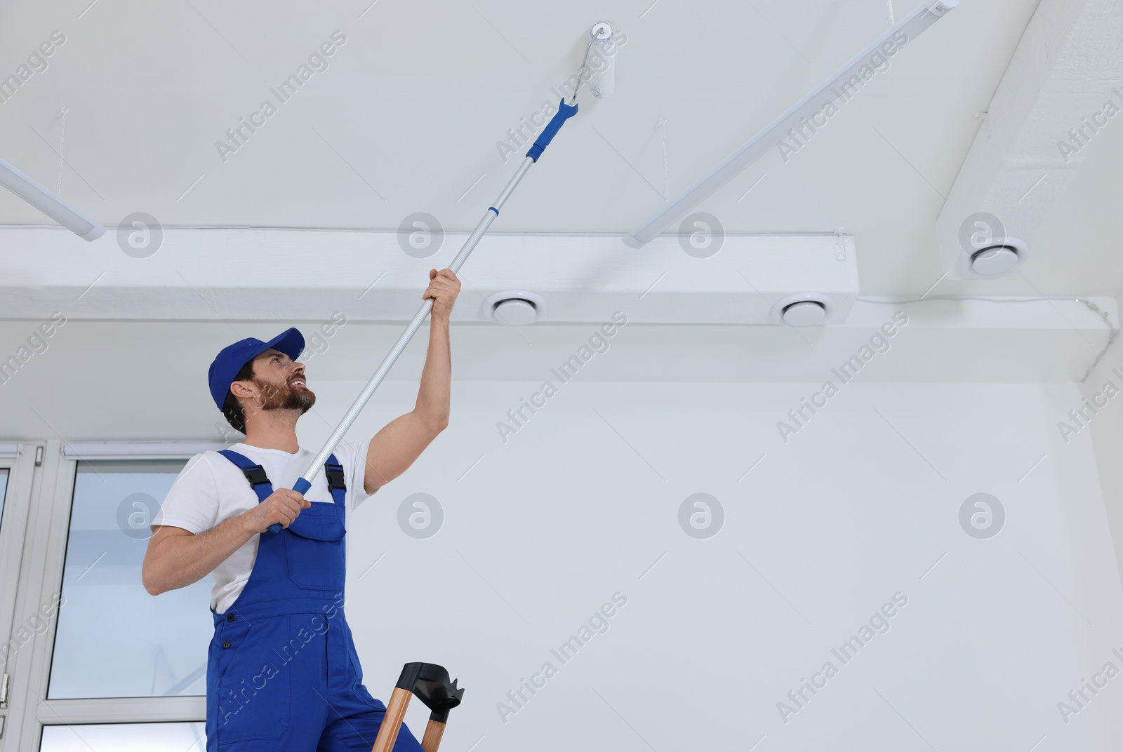 Photo of Handyman painting ceiling with roller on step ladder in room