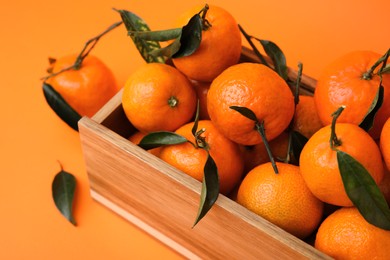Wooden crate with fresh ripe tangerines and leaves on orange table, closeup