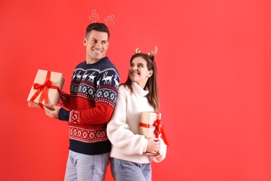 Photo of Beautiful happy couple in Christmas headbands and sweaters holding gifts on red background. Space for text