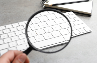 Photo of Woman holding magnifier glass near keyboard at light grey stone table, closeup. Find keywords concept