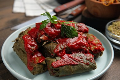 Photo of Plate of delicious stuffed grape leaves with tomato sauce and parsley on wooden table, closeup