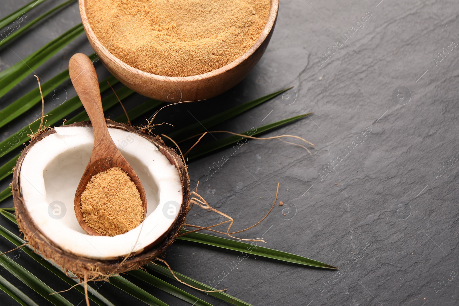 Photo of Spoon with coconut sugar, fruit, bowl and palm leaves on dark textured table, above view. Space for text