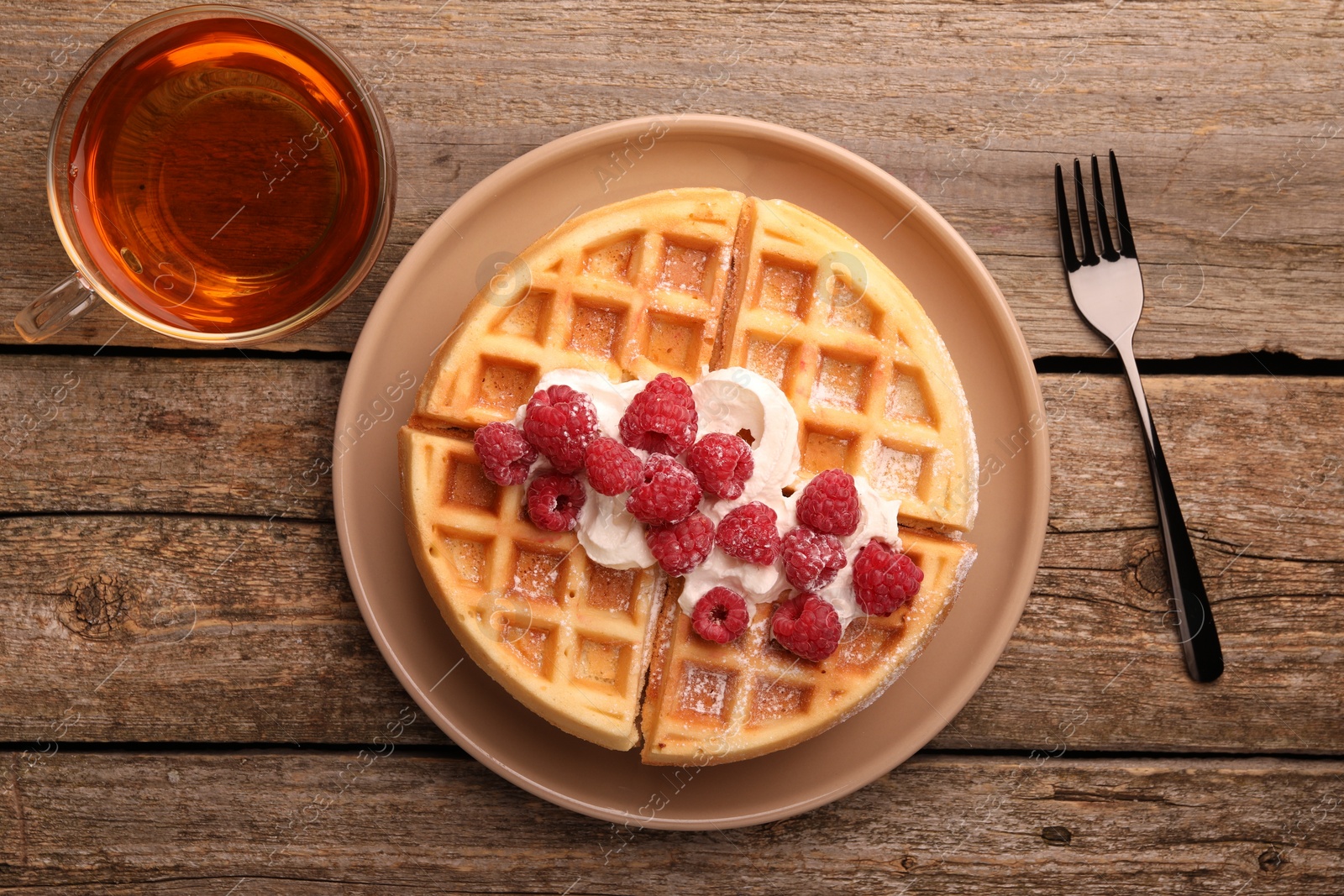 Photo of Tasty Belgian waffle with fresh raspberries, whipped cream, cup of tea and fork on wooden table, flat lay