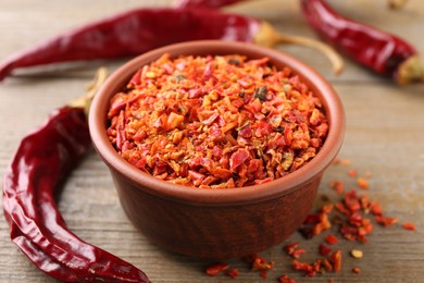 Photo of Aromatic spice. Red chili pepper flakes in bowl and pods on wooden table, closeup