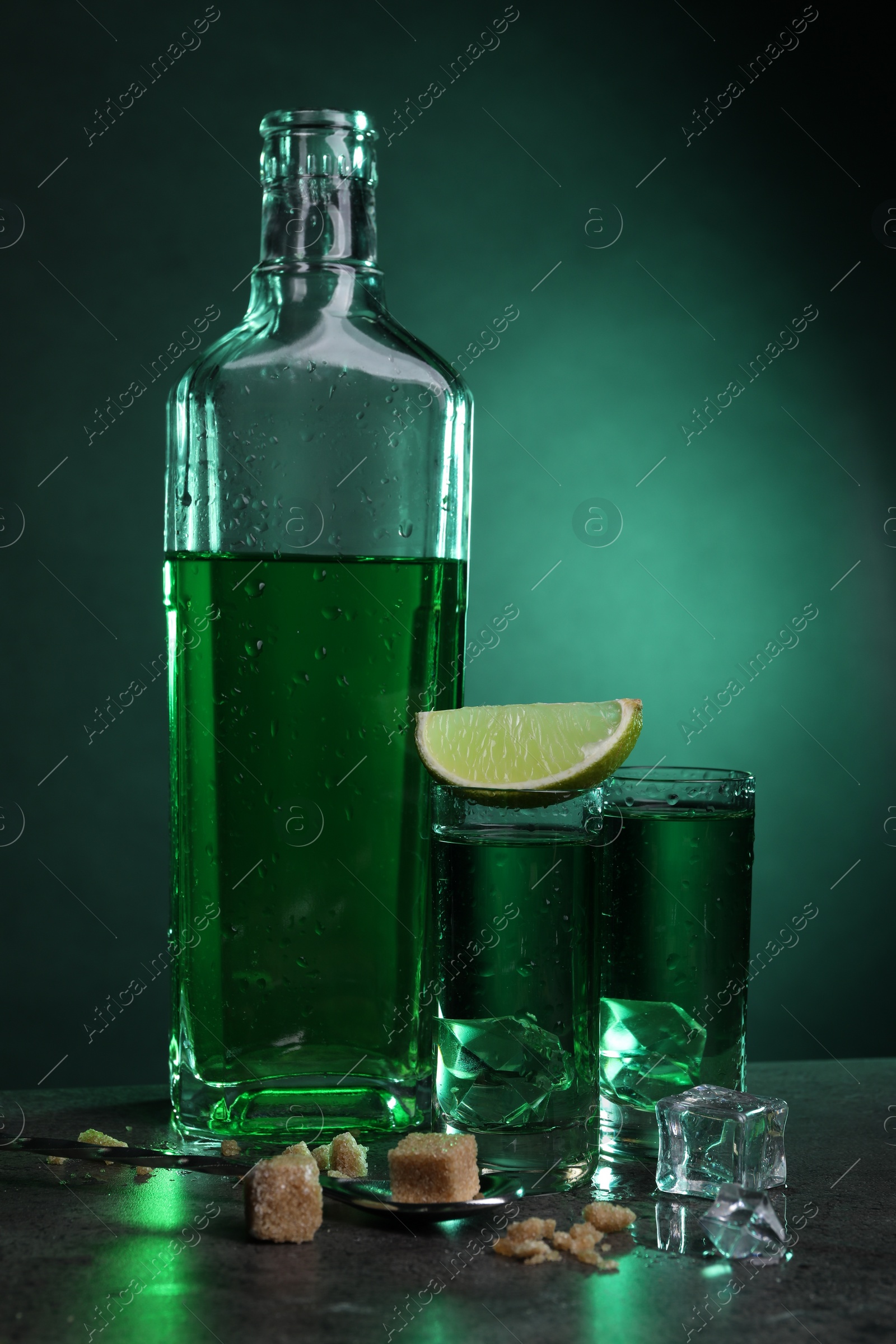 Photo of Absinthe in shot glasses with ice cubes, lime, brown sugar and spoon on gray table against green background. Alcoholic drink