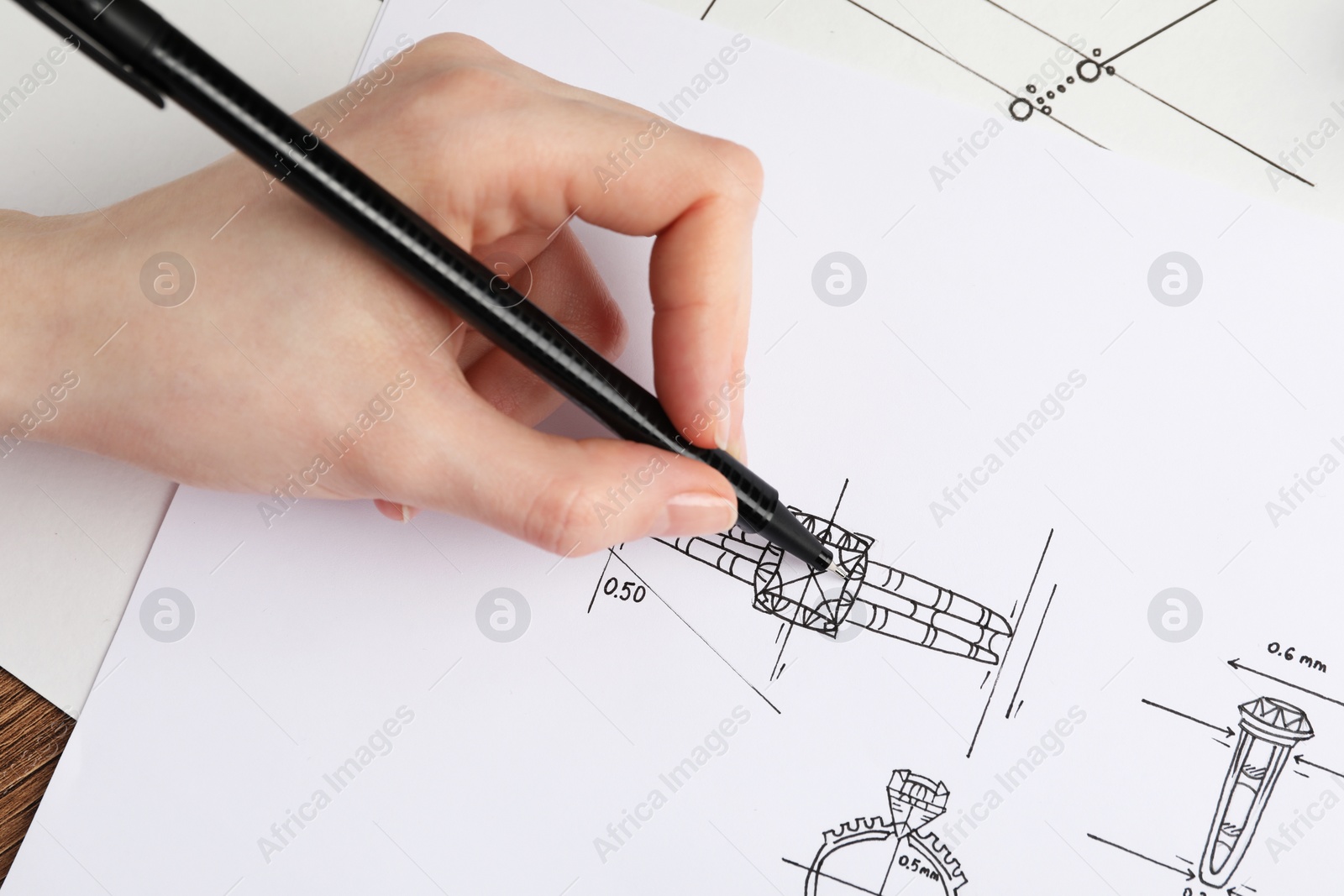 Photo of Jeweler drawing sketch of elegant ring on paper at wooden table, closeup