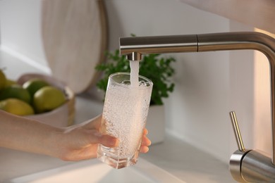 Woman filling glass with tap water from faucet in kitchen, closeup