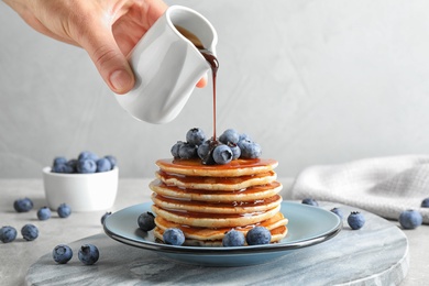 Photo of Woman pouring chocolate syrup onto fresh pancakes with blueberries at grey table, closeup