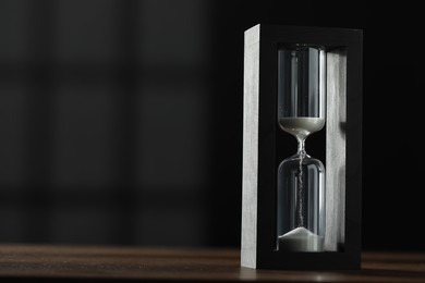 Photo of Hourglass with flowing sand on wooden table against black background, space for text