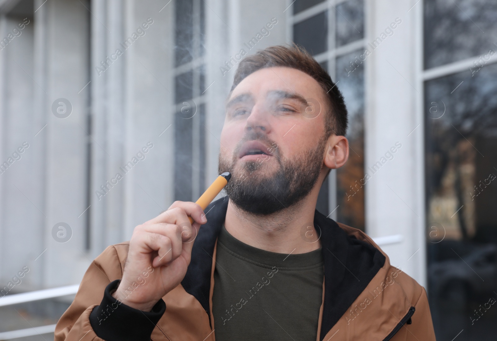 Photo of Handsome young man using disposable electronic cigarette outdoors