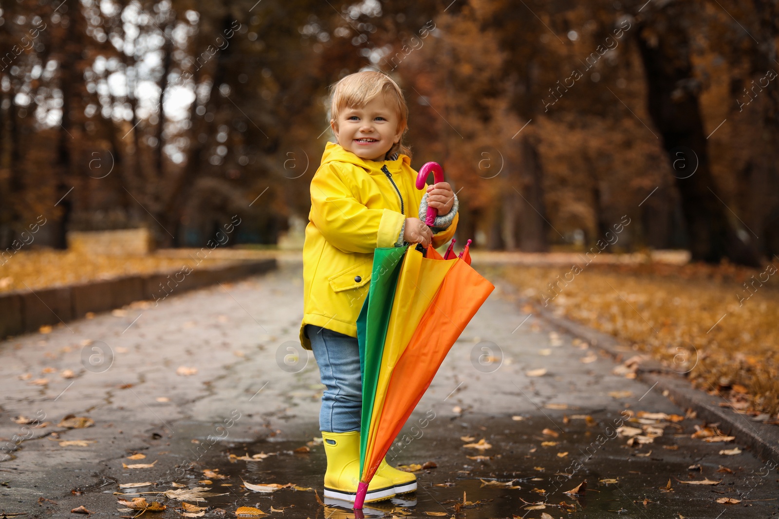 Photo of Cute little girl with colorful umbrella standing in puddle outdoors