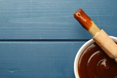 Photo of Tasty barbeque sauce in bowl and brush on blue wooden table, top view. Space for text