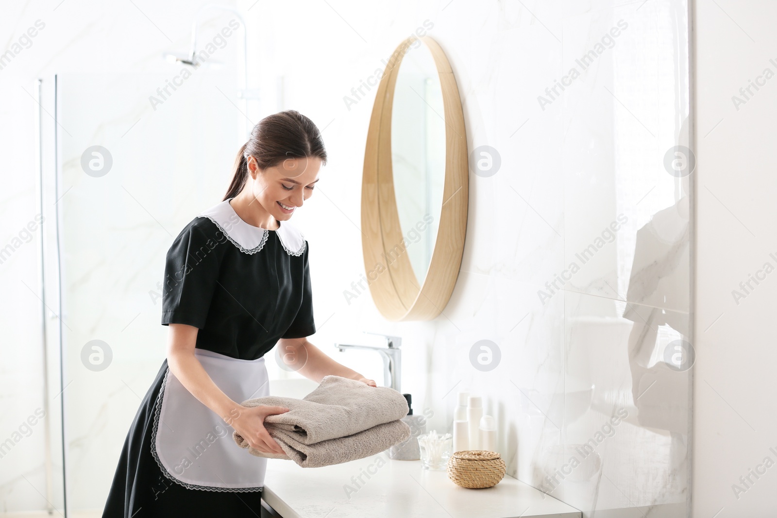 Photo of Young chambermaid putting stack of fresh towels on countertop in bathroom