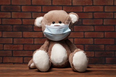Cute teddy bear with medical mask on wooden table near brick wall