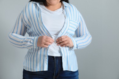 Overweight woman trying to button up tight shirt on light grey background, closeup