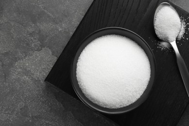 Photo of Granulated sugar in bowl and spoon on grey textured table, top view. Space for text