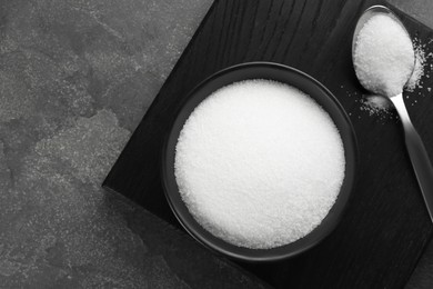 Granulated sugar in bowl and spoon on grey textured table, top view. Space for text