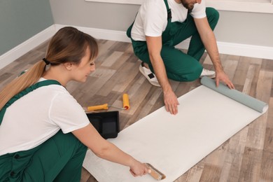 Photo of Workers applying glue onto wall paper sheet on floor indoors