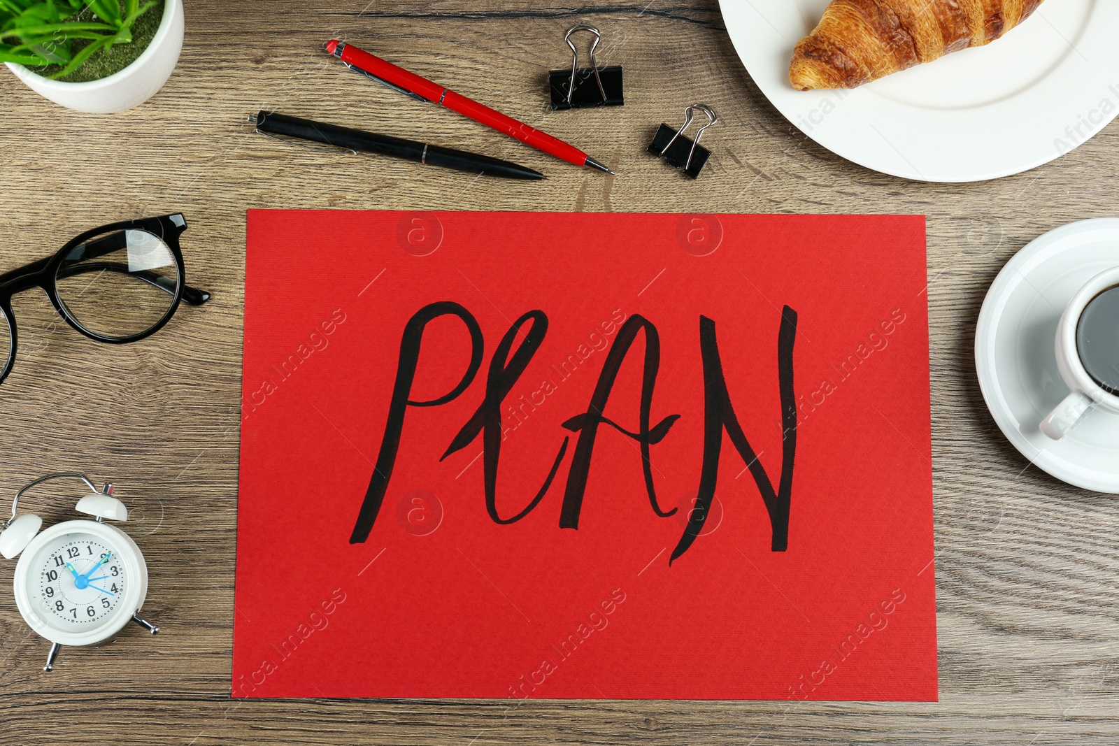 Photo of Paper with word Plan, stationery, alarm clock, croissant and coffee on wooden table, flat lay