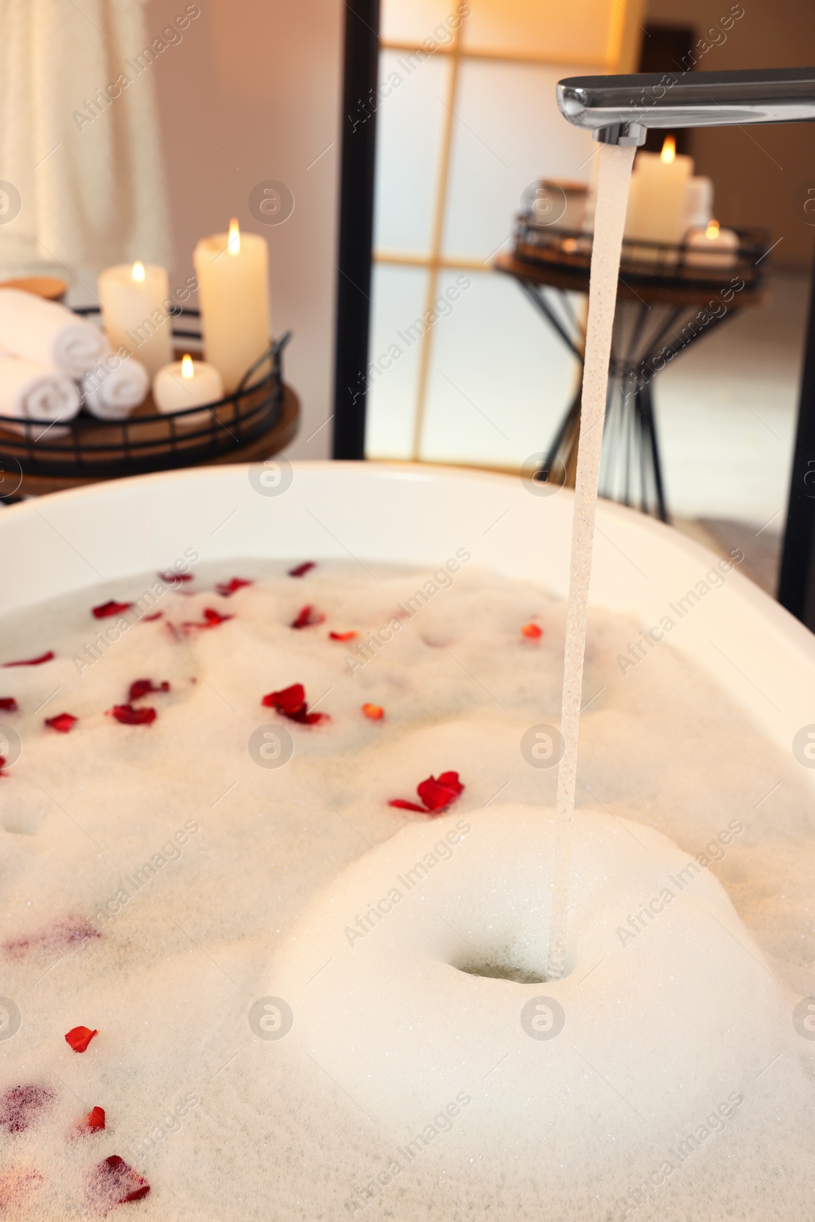 Photo of Water pouring into bath tub with foam and red rose petals in bathroom