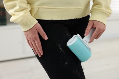 Photo of Woman with lint roller removing pet hair from black trousers indoors, closeup