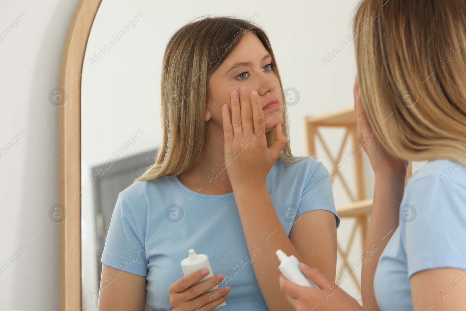 Photo of Sleep deprived young woman covering up dark circles with concealer near mirror at home