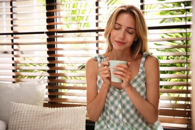 Photo of Beautiful young woman with cup of drink near window at home