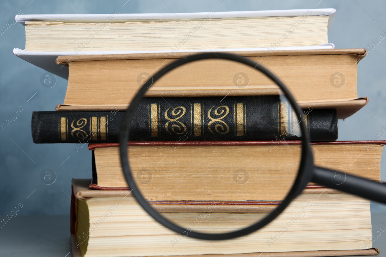 Photo of Looking through magnifying glass at stack of vintage books on grey table, closeup. Search concept