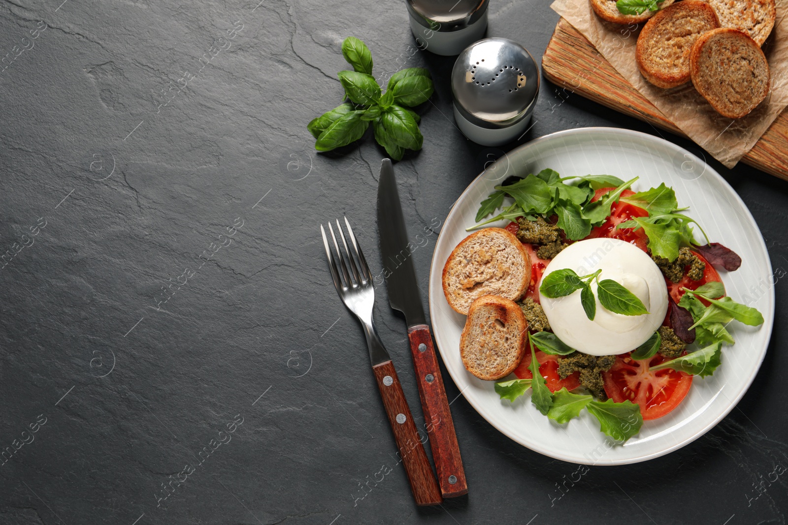 Photo of Delicious burrata salad with tomatoes, arugula and pesto sauce served on black table, flat lay. Space for text