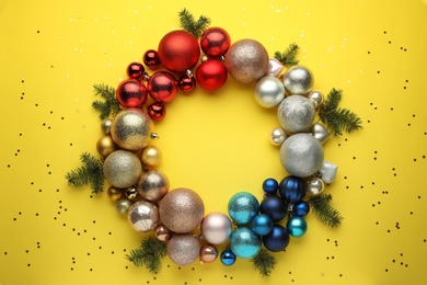 Bright festive wreath made of Christmas balls and fir branches on yellow background, top view. Space for text