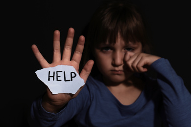 Photo of Crying little girl with sign HELP near black wall, focus on hand. Domestic violence concept