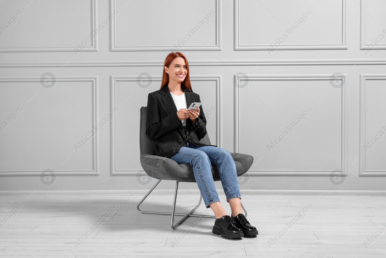 Photo of Happy young woman with smartphone sitting in armchair near light grey wall indoors